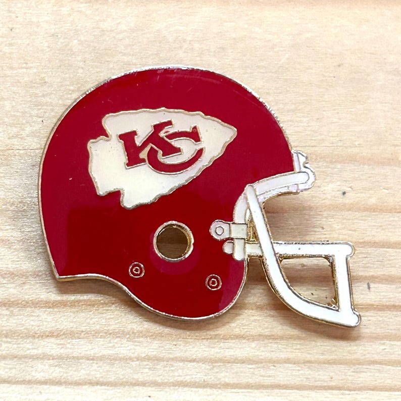May include: A red and white enamel pin in the shape of a football helmet. The helmet features the team's logo in white on the side. The pin has a gold-colored outline and a pin back.