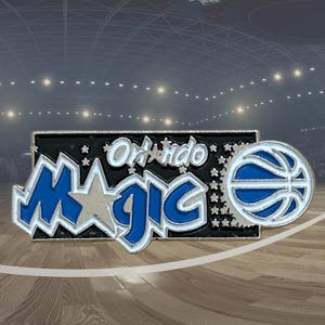 May include: A pin featuring the Orlando Magic logo. The pin has a black background with the words "Orlando Magic" in white and blue. A basketball is on the right side of the pin. The pin is likely a collectible item for basketball fans.