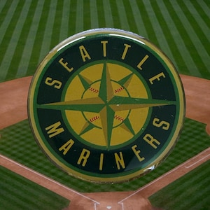 Puede incluir: Un pin redondo con el logotipo de los Marineros de Seattle. El pin presenta un diseño de rosa de los vientos en amarillo y verde, con el nombre del equipo en letras verdes. El fondo muestra un campo de béisbol.