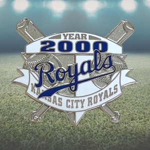 May include: A silver-colored pin featuring the text "YEAR 2000 Royals Kansas City Royals". The pin has a baseball theme with crossed bats and baseballs. The text is in blue and white.