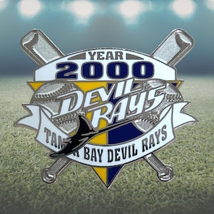 May include: A silver and enamel pin featuring the words "Year 2000" above "Devil Rays" and "Tampa Bay Devil Rays". The pin includes baseball bats, a baseball, and a stingray design, with blue, yellow, and white accents.