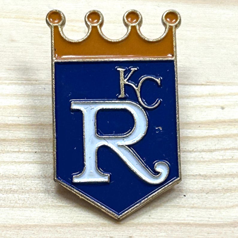 May include: A gold-coloured metal pin in the shape of a shield with a crown at the top. The shield is navy blue with a white "R" and "KC" in the upper right corner. The crown is orange.