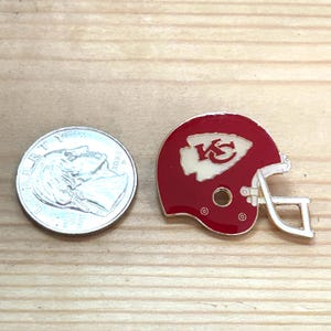 May include: A red and white enamel pin in the shape of a football helmet, featuring the Kansas City Chiefs logo. The pin is next to a US dime coin. The helmet has a white logo and a silver facemask.