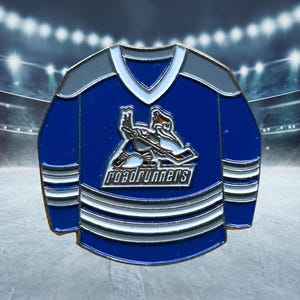 May include: A blue and gray hockey jersey pin featuring the Roadrunners logo. The pin has a white collar and stripes, with a hockey player graphic on the front. The background shows a hockey rink.