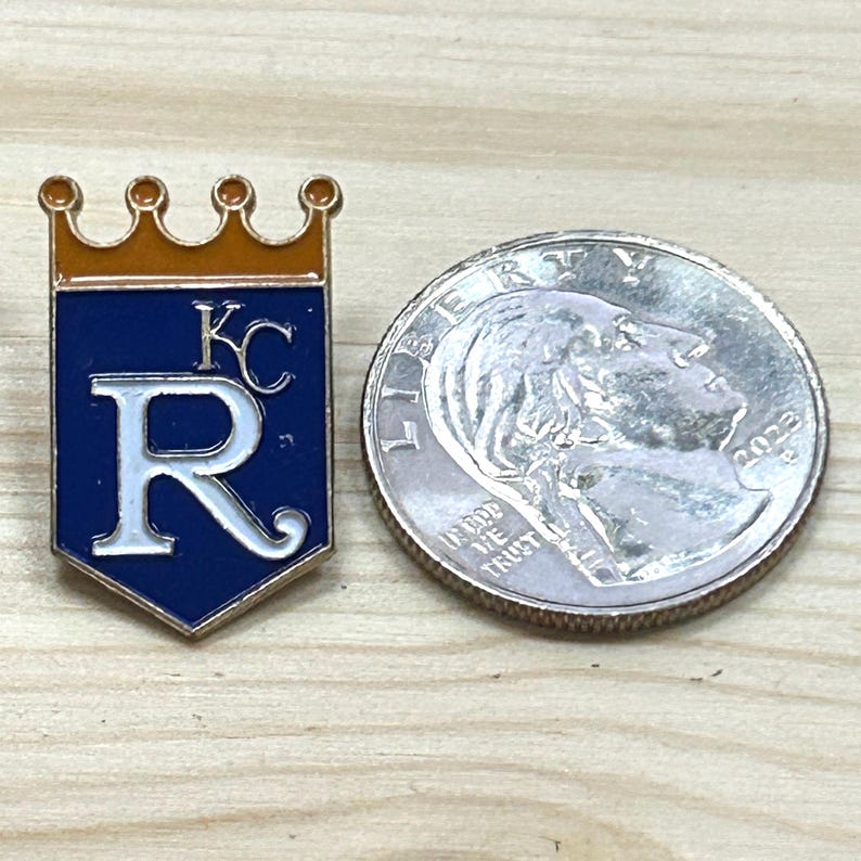 May include: A rectangular pin featuring a blue background with a white "R" and "KC" lettering, and a gold crown. A coin is next to the pin. The pin is likely a team logo or emblem.