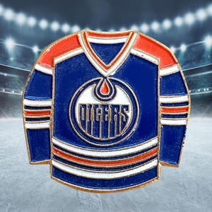 Op de afbeelding: Een blauw, oranje en witte emaille pin in de vorm van een hockeytrui. De trui heeft het woord "OILERS" in witte letters op een cirkelvormig embleem. De trui heeft oranje schouders en witte strepen.