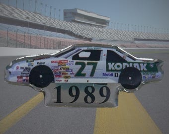 May include: A metallic pin shaped like a race car featuring the number 27, the text Kodiak, Mobil 1, Pontiac, and 1989. The car body is white with green accents and numerous small sponsor logos along the side panels and black wheels.
