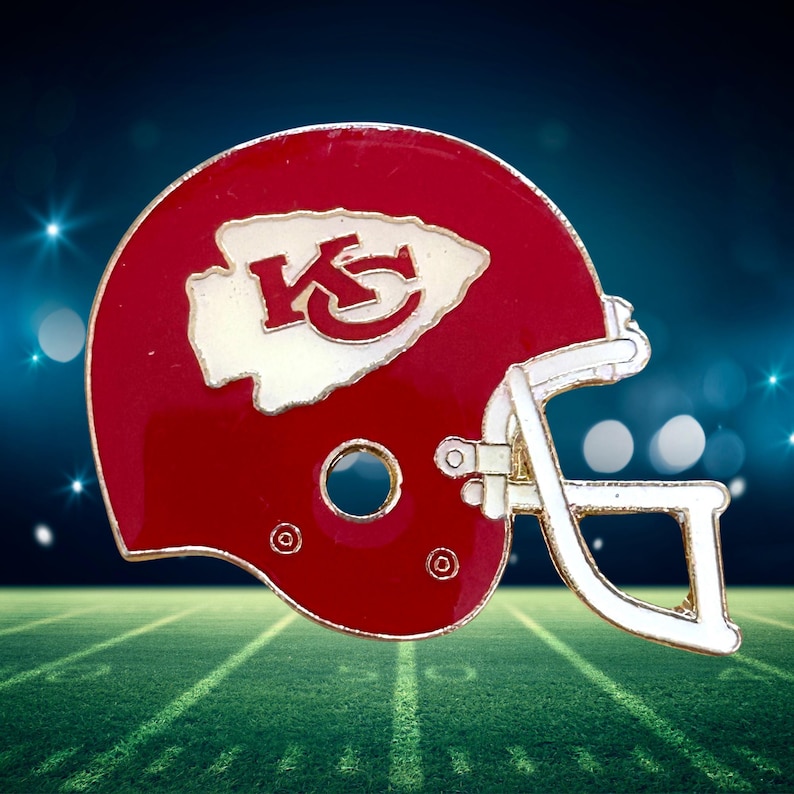 May include: A red and white football helmet pin featuring the Kansas City Chiefs logo. The helmet has a red shell with a white facemask and the team's logo prominently displayed on the side.