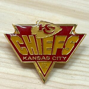 May include: A gold-toned metal lapel pin shaped like a shield with red enamel accents. The pin features the text KC, CHIEFS, KANSAS CITY, and NFL in gold lettering. The design is triangular with a pointed bottom and a jagged top edge.