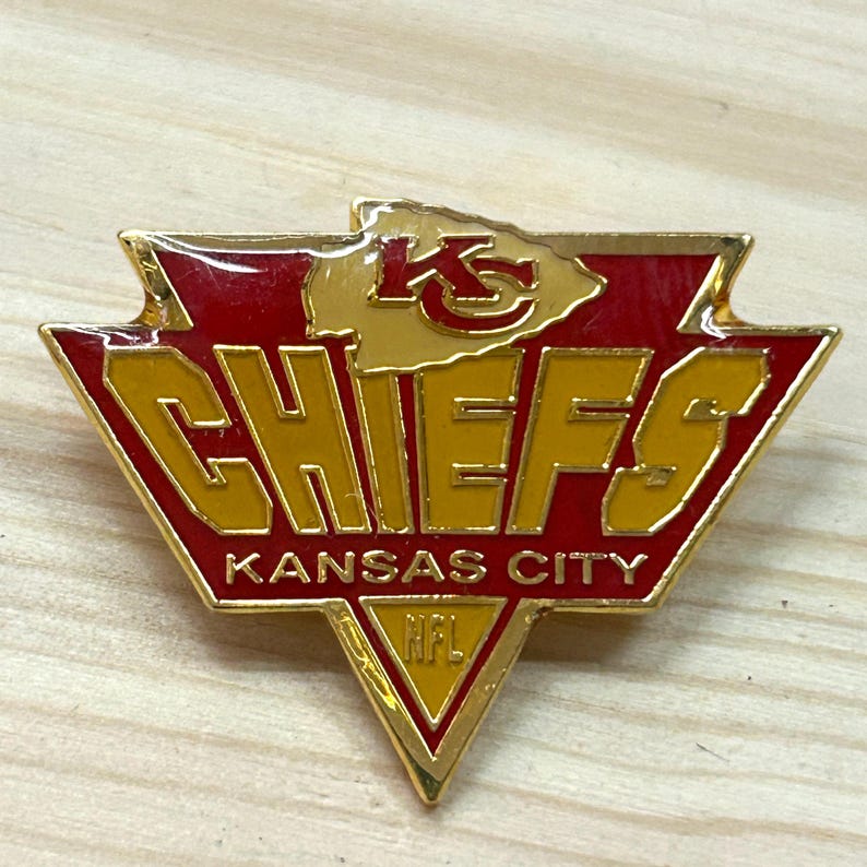 May include: A gold-toned metal lapel pin shaped like a shield with red enamel accents. The pin features the text KC, CHIEFS, KANSAS CITY, and NFL in gold lettering. The design is triangular with a pointed bottom and a jagged top edge.