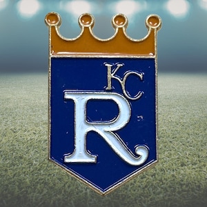 May include: A gold crown sits above a blue pennant-shaped pin. The pin has a white letter "R" and "KC" in white, with a gold border. This pin is likely a sports team emblem.