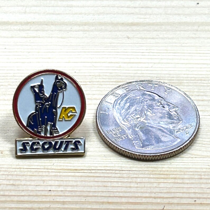 Puede incluir: Un pin de Scouts vintage con un dise&ntilde;o azul y blanco que presenta dos animales y las letras "KC" dentro de un c&iacute;rculo rojo. La palabra "SCOUTS" se muestra debajo del c&iacute;rculo. Una moneda de veinticinco centavos estadounidense est&aacute; al lado del pin.