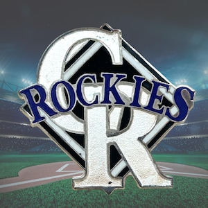 May include: A metal lapel pin featuring the Colorado Rockies baseball team logo. The design consists of a silver letter C and R intertwined with the word ROCKIES in blue lettering across the center, set against a black diamond background.
