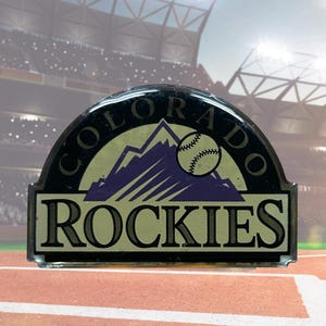 May include: A black and silver Colorado Rockies baseball team emblem. The emblem features the words "COLORADO" and "ROCKIES" with a baseball and mountain range graphic. The background shows a baseball field.