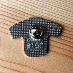 Puede incluir: Un pin de metal plateado con forma de camiseta de f&uacute;tbol con el logotipo de la NFL y el texto "Officially Licensed Product" y "STABLENE INC" en la parte posterior.