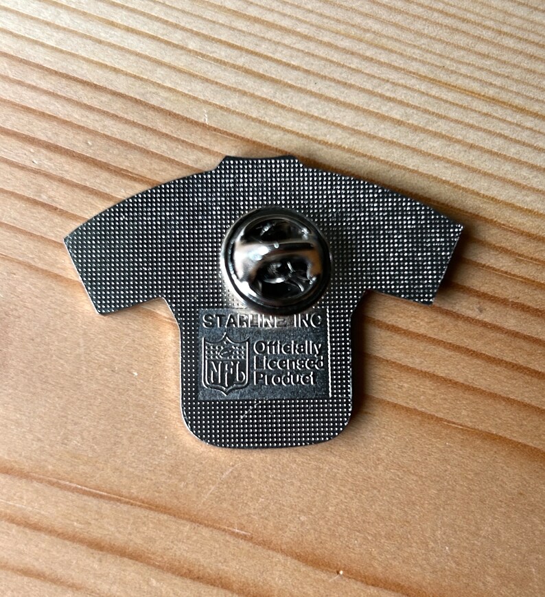 Puede incluir: Un pin de metal plateado con forma de camiseta de f&uacute;tbol con el logotipo de la NFL y el texto "Officially Licensed Product" y "STABLENE INC" en la parte posterior.
