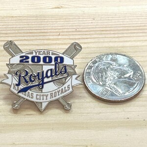 May include: A silver-colored pin featuring the Kansas City Royals logo with the text "YEAR 2000 Royals" in blue and white. Two crossed baseball bats are behind the logo. A quarter is next to the pin.