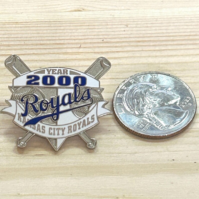 May include: A silver-colored pin featuring the Kansas City Royals logo with the text "YEAR 2000 Royals" in blue and white. Two crossed baseball bats are behind the logo. A quarter is next to the pin.