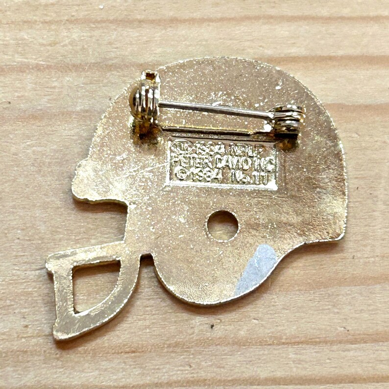 May include: A gold-colored football helmet pin with a pin back. The pin is in the shape of a football helmet with a face mask. The back of the pin has the text "&copy;1984 NFL PETER DAVID INC."