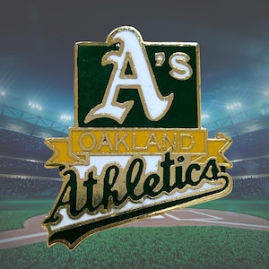 May include: A metal lapel pin featuring the Oakland Athletics logo. The design includes a white letter A's on a dark green square, a yellow banner with the word OAKLAND in gold, and the word Athletics in dark green script on a white background.