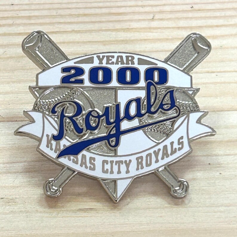 May include: A silver-colored pin featuring the Kansas City Royals logo. The pin has a white background with blue text that reads "YEAR 2000 Royals" and "KANSAS CITY ROYALS". Two crossed baseball bats are behind the logo.