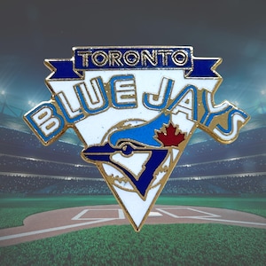 May include: A metal enamel pin featuring the Toronto Blue Jays logo. The design includes a white triangle background, a blue jay bird head silhouette, a red maple leaf, and the text TORONTO BLUE JAYS in blue lettering with gold trim.