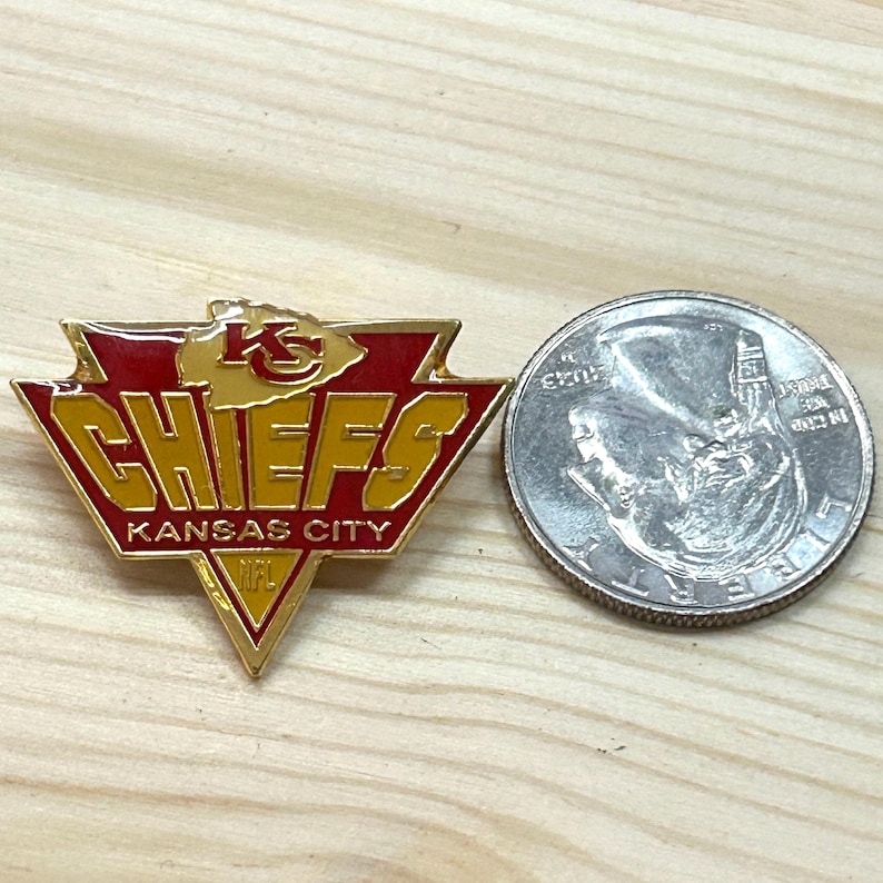 May include: A triangular Kansas City Chiefs enamel pin featuring red and gold colors with the team logo, the word CHIEFS, and the text KANSAS CITY NFL. The pin is shown next to a United States quarter coin for scale, which has a diameter of 0.955 inches.