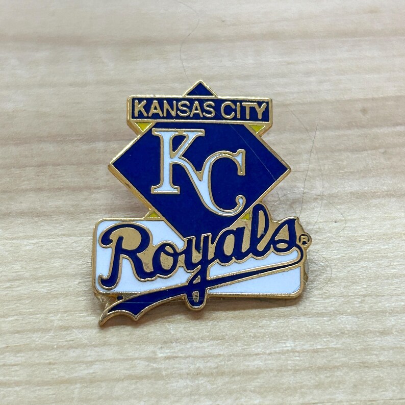 May include: A gold-colored pin with a blue diamond shape with the text "KANSAS CITY" and "KC" in white. Below the diamond is a white banner with the text "Royals" in gold.