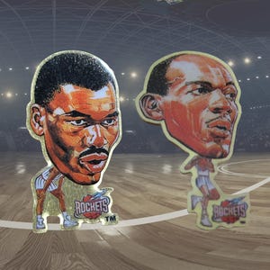 May include: Two basketball player caricatures with gold trim. The players are depicted in action poses, with the Houston Rockets logo at the bottom. The background shows a basketball court and arena.