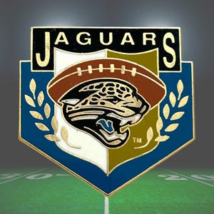 May include: A metal enamel pin shaped like a shield with a blue border, featuring the text JAGUARS at the top. The center displays a brown football above a black and gold jaguar head logo, flanked by gold laurel leaves on a white and gold background.