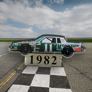 May include: A green and white race car with the number 11 and the words "Mountain Dew" on the side. The car is on a checkered flag finish line and the year 1982 is printed below the car.