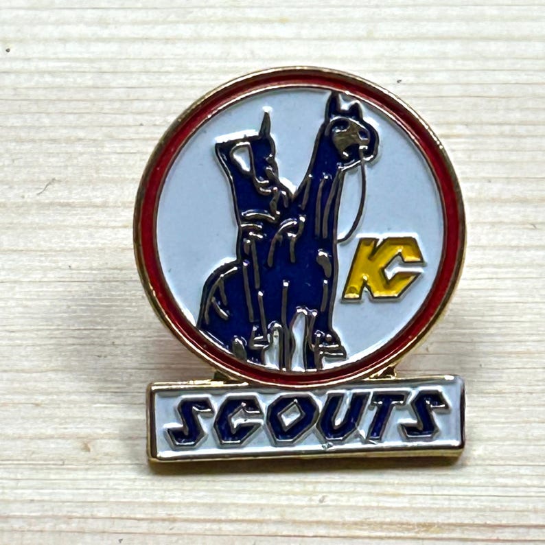 Puede incluir: Un pin vintage de los Scouts con borde dorado. El pin presenta un dise&ntilde;o azul y blanco de dos animales y las letras "KC" en amarillo. La palabra "SCOUTS" se muestra en letras blancas sobre un fondo dorado.