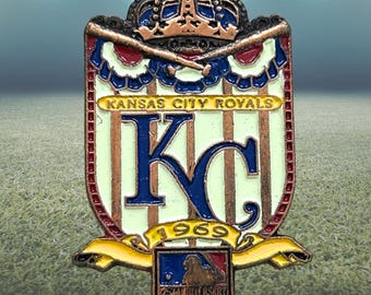 Könnte beinhalten: Ein Ansteckpin des Baseballteams Kansas City Royals. Der Pin zeigt das Logo des Teams, ein stilisiertes "KC" in Blau, mit "Kansas City Royals" darüber. Der Pin enthält auch das Jahr "1969" und ein Baseball-Logo.