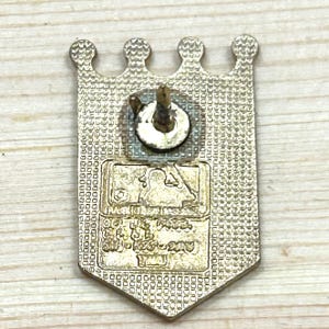 May include: A gold-coloured metal pin with a crown-shaped top and a rectangular bottom. The pin features the MLB logo and text. The pin has a textured surface and a pin back.
