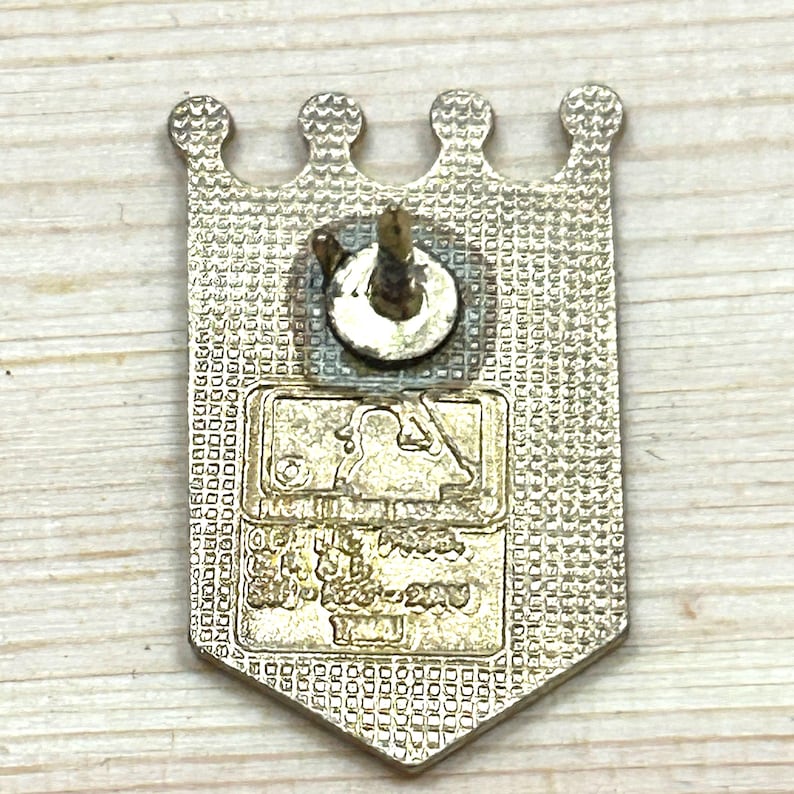 May include: A gold-coloured metal pin with a crown-shaped top and a rectangular bottom. The pin features the MLB logo and text. The pin has a textured surface and a pin back.