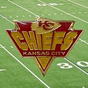 May include: A metallic Kansas City Chiefs pin featuring a red and gold arrowhead logo with the text KC, CHIEFS, KANSAS CITY, and NFL. The design is shaped like a triangular shield with a pointed bottom and a top arrowhead silhouette.