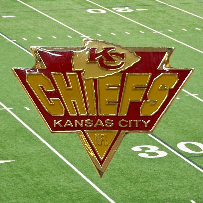 May include: A metallic Kansas City Chiefs pin featuring a red and gold arrowhead logo with the text KC, CHIEFS, KANSAS CITY, and NFL. The design is shaped like a triangular shield with a pointed bottom and a top arrowhead silhouette.