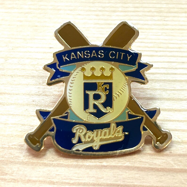 May include: A Kansas City Royals baseball pin with a gold border. The pin features crossed baseball bats, a baseball, and banners with the words "Kansas City" and "Royals". The baseball has a crown and the letter "R".