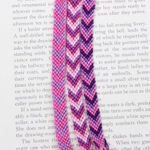 May include: Four handmade friendship bracelets in shades of pink, purple, and white, arranged on a book page. Each bracelet features a chevron pattern, showcasing a blend of colors. The bracelets are made of woven thread.