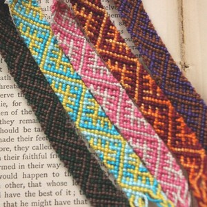 May include: Close-up of six colorful friendship bracelets with intricate woven patterns. The bracelets feature a variety of colors, including brown, blue, pink, orange, and purple. The bracelets are lying on top of an open book.