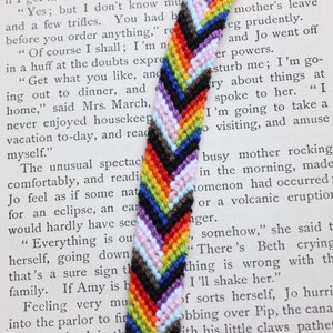 May include: A multi-colored chevron friendship bracelet with a rainbow pattern. The bracelet is made of woven thread and has a black, brown, white, light blue, purple, yellow, orange, and red color scheme.