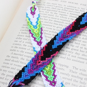 May include: Two colorful friendship bracelets with a chevron pattern. One bracelet is white with green and purple accents, the other is black with blue, pink, and purple accents.