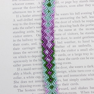 May include: A close-up of a purple, green, and white friendship bracelet with a chevron pattern. The bracelet is made with embroidery floss and is tied in a knot at the end.