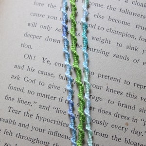 May include: Four colorful braided cords in shades of blue, green, and white. The cords are twisted together and are lying on a page of a book.