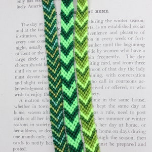 May include: Three green, yellow, and blue chevron friendship bracelets with a white background. The bracelets are made with a woven technique.