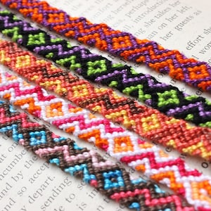 May include: A set of seven colorful friendship bracelets with a chevron pattern. The bracelets are made with different colored threads and are arranged in a row on a book.