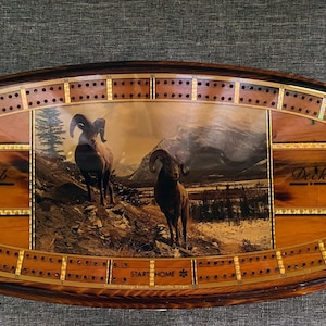 May include: An oval wooden cribbage board featuring a central graphic of two bighorn sheep in a mountain landscape. The board includes inlaid scoring tracks with holes, the text "Crib" and "Deck" on the sides, and "START HOME" at the bottom.