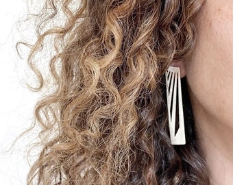 May include: Silver-colored rectangular earrings with a geometric design. The earrings have a series of vertical lines cut into the metal, creating a striped pattern. The earrings are hanging from a person's ear, with curly brown hair in the background.