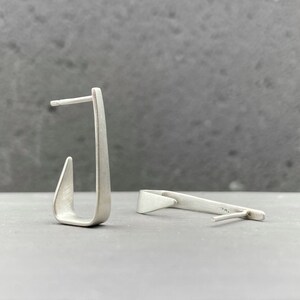 May include: A pair of silver earrings with a unique, asymmetrical design. The earrings are made of a smooth, brushed metal and have a simple, modern aesthetic.