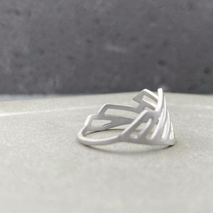 Pode incluir: Um anel de prata com um design geom&eacute;trico. O anel &eacute; feito de uma &uacute;nica pe&ccedil;a de metal e tem um design delicado e vazado.